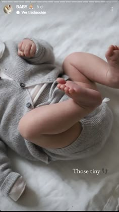 a baby laying on top of a bed wearing a gray sweater and white pants with one foot in the air