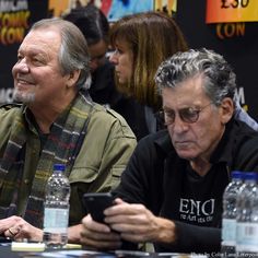 two men sitting next to each other at a table with water bottles and cell phones