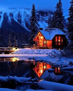 a cabin is lit up at night in the snow near a mountain lake and trees