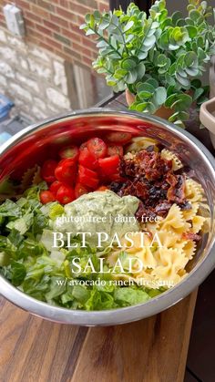 a bowl filled with pasta and vegetables on top of a wooden table next to a potted plant