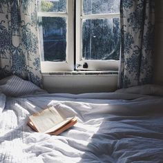 an open book sitting on top of a bed in front of a window with curtains