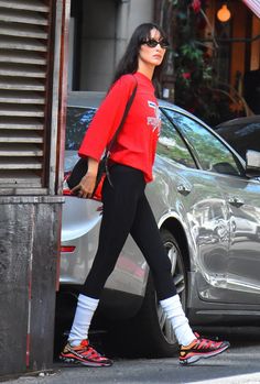 a woman in red shirt and black leggings walking down street next to car