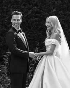 a bride and groom standing next to each other in front of some bushes holding hands