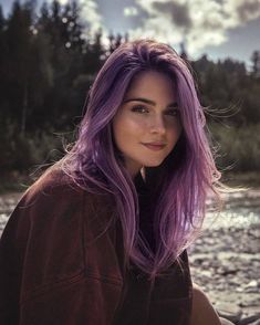 a woman with purple hair is sitting on the rocks in front of a body of water