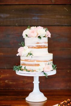 a three tiered cake with flowers on the top is sitting on a table in front of a wooden wall