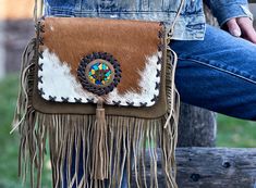 The Lakota Mountain Fringe Cowhide Crossbody Purse is a stylish and unique bag made of soft and durable suede leather and cowhide. The flap cover is made of cowhide and features a round ornament made, adding a touch of artistic flair to the bag. The bag also has decorative leather embroidery on the cover and fringes on the sides, adding texture and interest to the design. This bag is perfect for anyone looking for a fashionable and practical accessory to complete their outfit. NOTE: Each bag is Western Style Crossbody Shoulder Bag For Travel, Western Style Rectangular Bag With Adjustable Strap, Western Brown Satchel Shoulder Bag, Western Style Brown Satchel Bag, Rectangular Brown Bag For Rodeo, Western Brown Bags For Rodeo, Brown Rectangular Bag For Rodeo, Western Style Crossbody Bags For Western-themed Events, Western Style Brown Bags For Rodeo