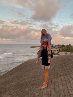 a woman carrying a child on her back while walking along the boardwalk near the ocean