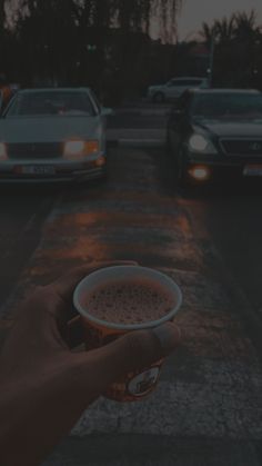 a hand holding a cup of coffee in front of parked cars on the side of a road