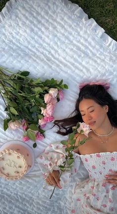 a woman laying on top of a white blanket next to flowers and a bowl of cake