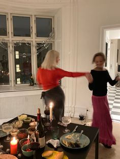 two women dancing around a table full of food and drinks