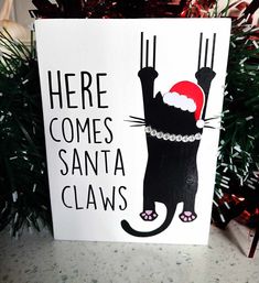 a christmas card with a black cat wearing a santa hat and saying here comes santa claws