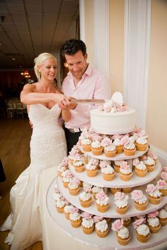 a bride and groom cutting their wedding cake with cupcakes in front of them