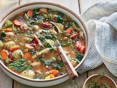 a bowl of soup with carrots, broccoli and other vegetables next to a wooden spoon