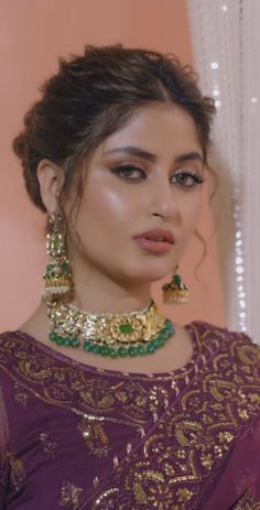 a woman in a purple dress with gold and green jewelry on her neck, looking at the camera