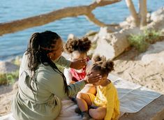 a woman and two children sitting on a blanket by the water with their arms around each other