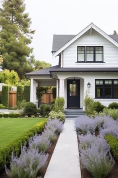 a white house with purple flowers in the front yard and bushes on either side of it