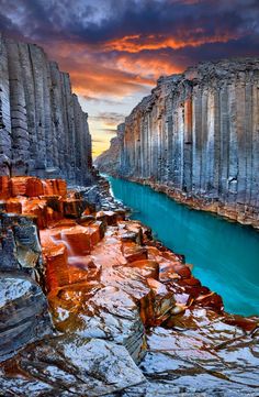 a river flowing through a canyon next to rocky cliffs under a colorful sky with clouds