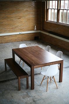 a wooden table with white chairs and a bench in an empty room next to a brick wall