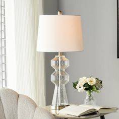 a table with a lamp, book and flowers on it in front of a window