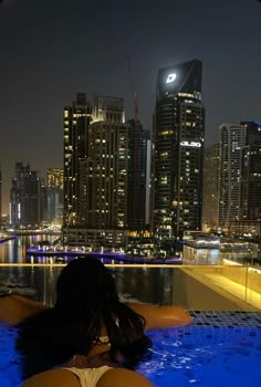 a woman sitting in the middle of a swimming pool with city lights in the background