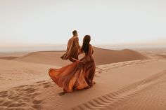 a man and woman walking across a sandy desert field in front of sand dunes at sunset