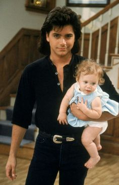 a man holding a baby in his arms while standing next to a stair case and wooden floors