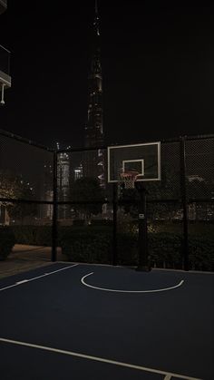 an empty basketball court in front of a cityscape at night with the lights on