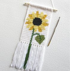 a cross stitch sunflower on a white wall hanging with a needle and thread next to it