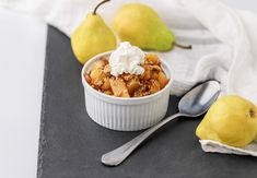 an apple crisp with whipped cream in a white dish next to two pears and a spoon