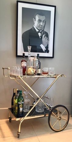 a bar cart with glasses and bottles on it in front of a framed photo above