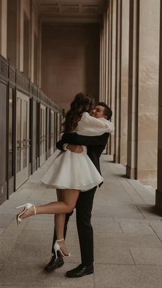 a man and woman are hugging on the sidewalk in front of some building pillars, one is wearing high heeled shoes