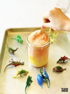 a hand is holding a spoon over a jar filled with toy dinosaurs on a tray