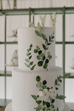 a three tiered white cake with green leaves and flowers on the top, sitting on a wooden table