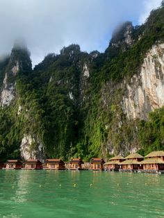 there are many huts in the water by the mountains and trees on the side of the mountain