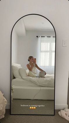 a woman sitting on top of a white bed in front of a mirror with her reflection