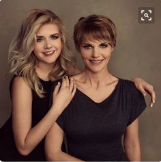 two beautiful women standing next to each other in front of a gray background with one holding her arm around the other's shoulder