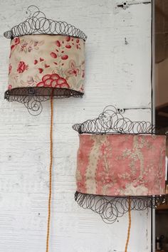 two lamps hanging on the side of a white wall with rusted iron wire and fabric coverings