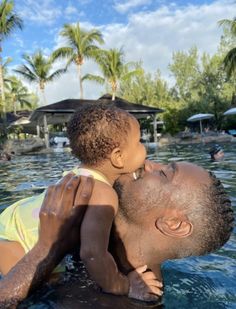 a man holding a small child in the water at a resort swimming pool with palm trees behind him