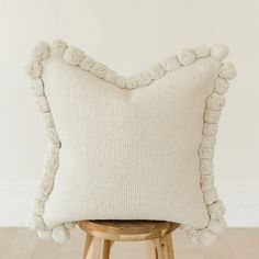 a white pillow with pom poms on it sitting on a stool in front of a wall