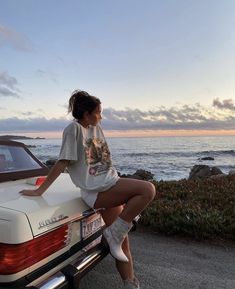 a woman sitting on the back of a white car near the ocean with her legs crossed