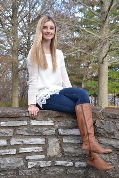 a young woman sitting on top of a stone wall wearing boots and smiling at the camera