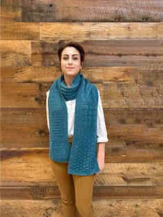 a woman standing in front of a wooden wall wearing a blue scarf and tan pants