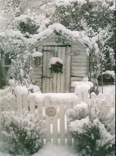 a white picket fence covered in snow next to a small wooden shed with a wreath on the door