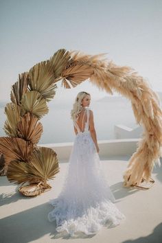 a woman in a wedding dress standing under an arch made out of palm leaves and feathers