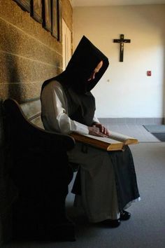 a nun sitting on a bench reading a book in front of a wall with a cross above her head