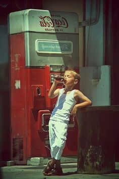 a young boy leaning against a post while talking on a cell phone in front of a coca cola machine