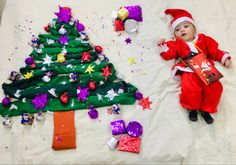 a baby laying next to a christmas tree with presents on the ground and confetti scattered around it