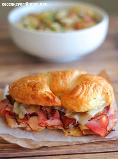 a croissant sandwich with meat and cheese on it next to a bowl of soup