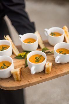 a wooden tray topped with cups of soup next to toasted bread on top of a table