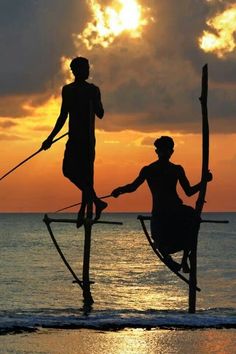 two people are standing on stilts in the water at sunset, with one holding an oar